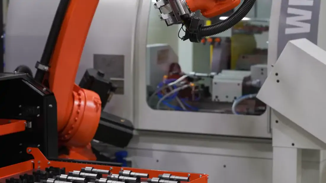 A KUKA robot reaching into an open machine to seat a shaft in the chuck